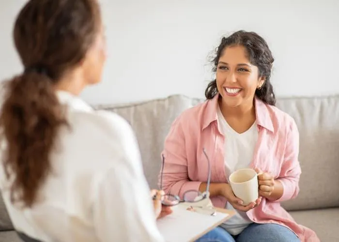 A woman wearing a pink shirt sits on a couch and smiles while holding a cup of coffee. She is talking to another woman sitting next to her, who is wearing a white shirt.