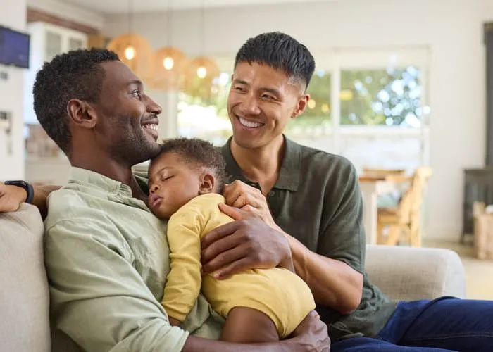 Same Sex Male Couple Or Friends Cuddling Sleeping Baby Daughter On Sofa At Home Together