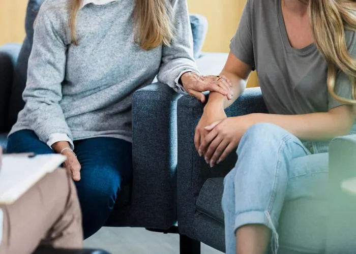 An unrecognizable mother touches her daughter's arm while talking with a therapist during a counseling session.