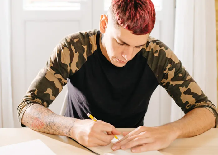 Young stylish man sitting and writing with pen in notepad at workplace.